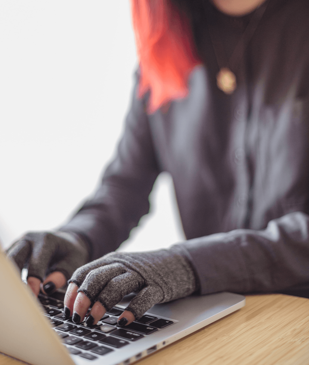 Una persona asiática con discapacidad y de género fluido que tiene puestos unos guantes de compresión escribe en una computadora portátil. Las manos y el teclado son el punto focal.