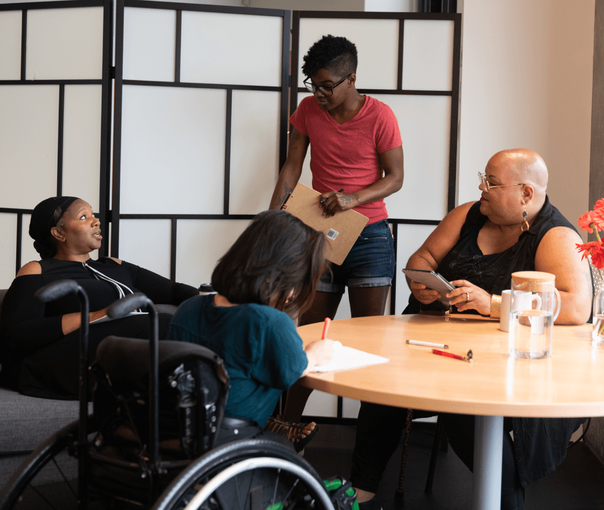 Cuatro personas discapacitadas de color están reunidas en torno a una mesa. Una mujer negra sentada en un sofá habla con expresión neutral mientras las otras tres escuchan (una persona del sur de Asia que toma notas sentada en una silla de ruedas; una persona negra no binaria sentada en una silla, que sostiene una tableta y tiene un bastón, y una persona negra no binaria que está de pie y lleva un portapapeles en la mano).